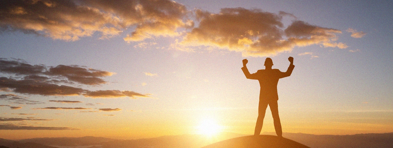 A man on top of a hill, facing the sunset, striking a superhero pose as if he has succeeded.